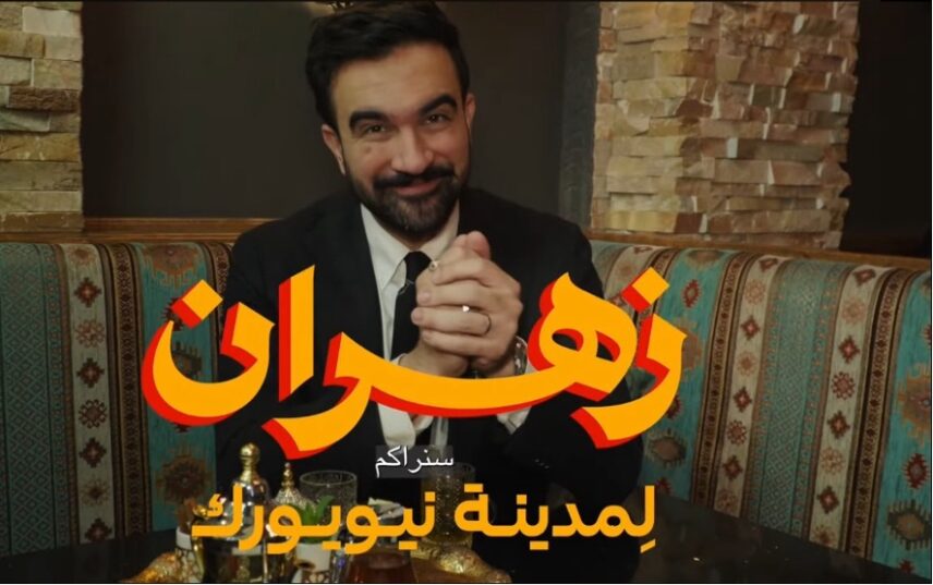 Zohran Mamdani, a man in his thirties with black hair, a mustache, and small beard is sitting in an upholstered booth with decorative green, gold, and white stripes at a Levantine restaurant. He is wearing a black suit, white shirt, and black tie. On the table in front of him is a gold tray, which includes a silver tea pot, a small glass, and a white container with mint leaves. Floating in front of the image is Arabic text in orange font, which states: Zohran for the City of New York.