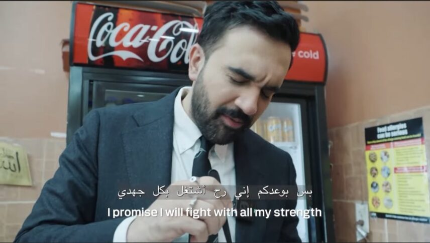 Zohran Mamdani, a Brown man in his thirties with black hair, a mustache, and small beard is eating a piece of an Arabic dessert, Nabulsi knafeh, while standing at Levantine bakery in New York City. He is wearing a black suit, white shirt, and black tie and behind him is a commercial beverage refrigerator with Coca Cola written on the top of it. Mamdani is making a face that shows he is enjoying the dessert.