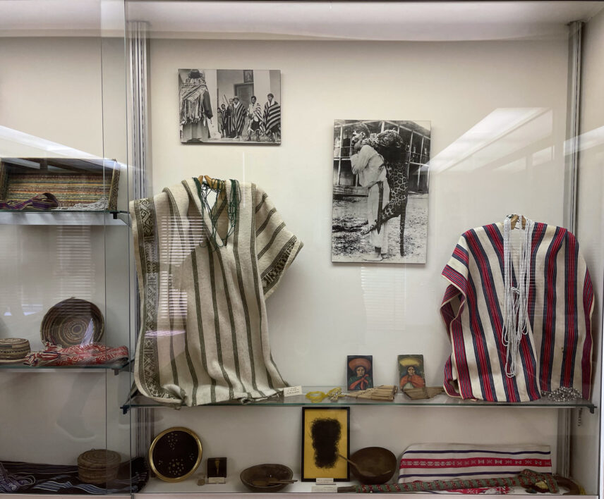 Museum vitrine displaying Kamëntšá and Inga textiles, baskets, ceremonial objects, musical instruments, and historical photographs.