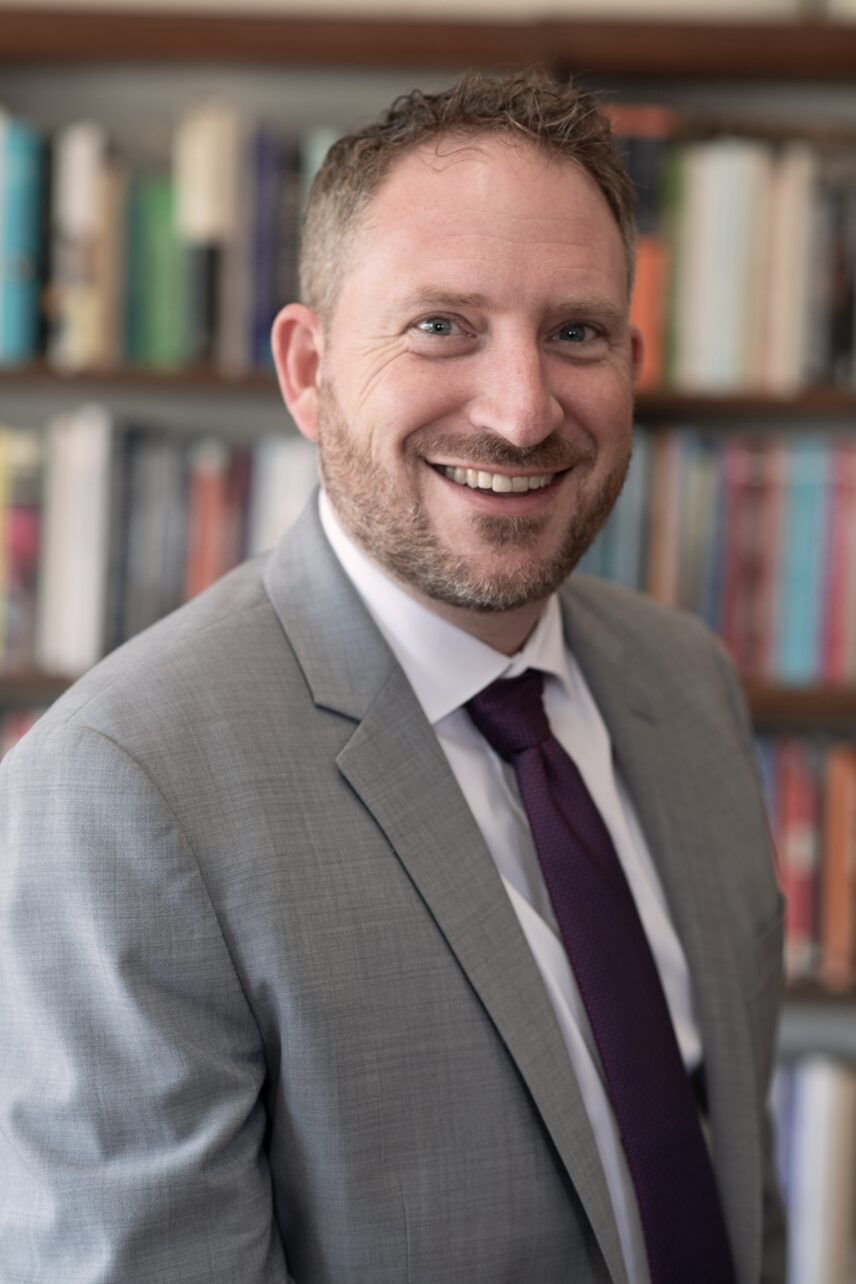 Michael A. Di Giovine dressed in a suit in his office in front of book shelf, smiling