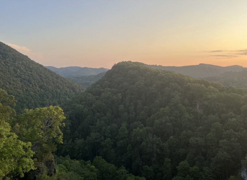 A sunset view of the Cumberland Mountains in Southwest Virginia during late summer. The mountains are full of greenery, and the sun has set behind them to paint the sky soft oranges and yellows.