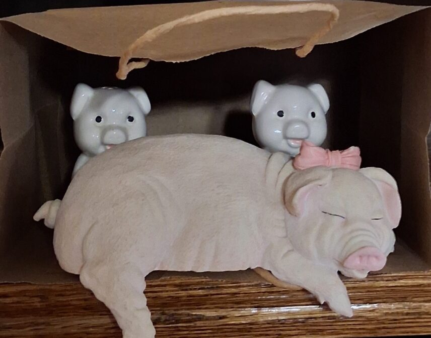 A “shelf-sitter” in the shape of a sleeping pink pig rests on a shelf. White salt and pepper shakers also shaped like pigs peek out from behind the shelf sitter. A brown paper bag is set up around the figurines to display “a pig in a poke.”
