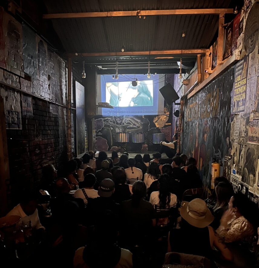 A large crowd of people seated in a narrow, dark room illuminated by a film projector