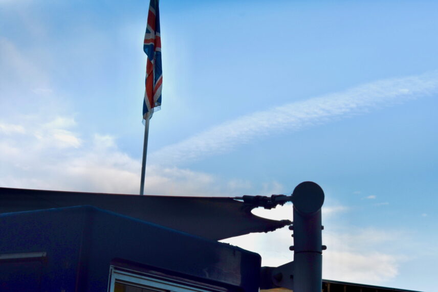 Close-up of the top edge of a dark blue building with a shade structure. A British Union Jack flag hangs from a thin white flagpole, backlit against a blue sky with light clouds.
