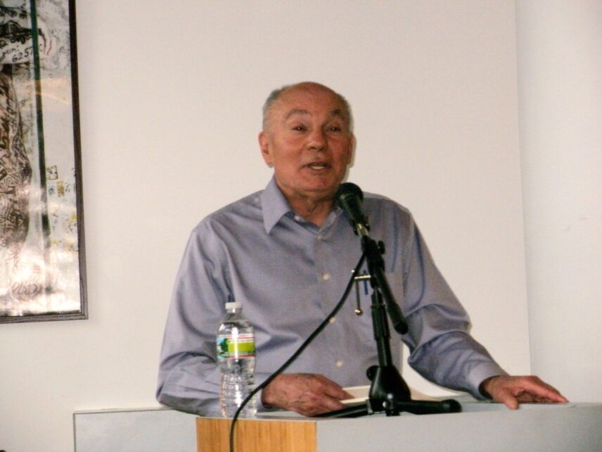 Renato Rosaldo in a button-down shirt speaking into a microphone while standing behind a podium with a bottle of water nearby.