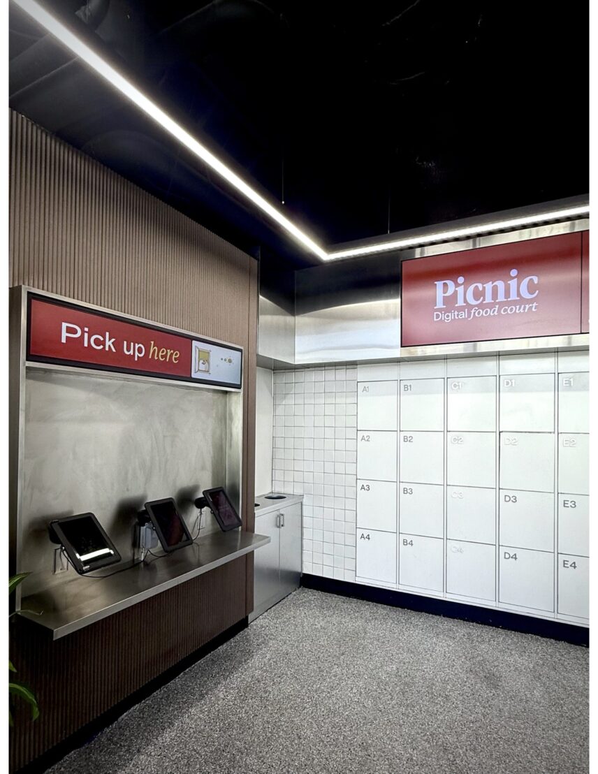An example of a pickup area in a local ghost kitchen. There are three tablets on the left, which a driver will use to open up one of the numbered/lettered lockers present along the wall.