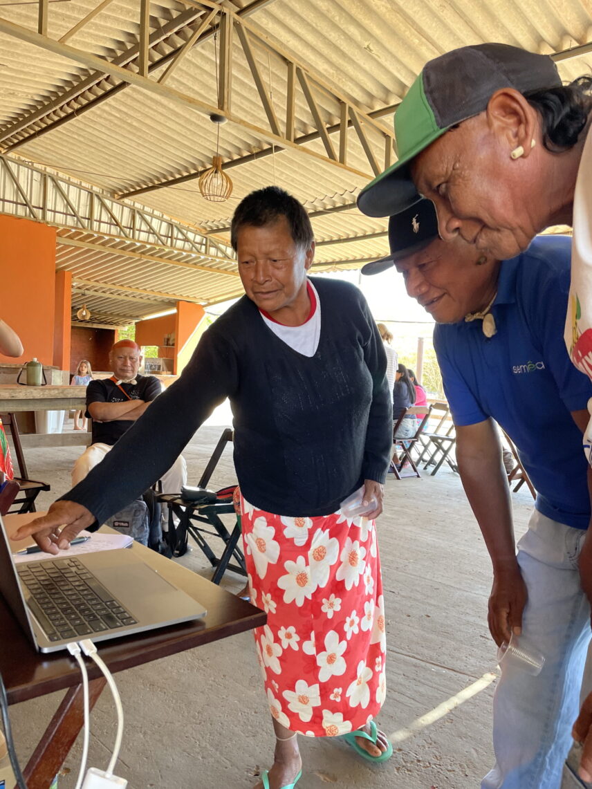 An A’uwẽ woman wearing a black sweater and a red floral skirt stands and points at a laptop screen while two A’uwẽ men wearing baseball caps look at the screen, smiling.