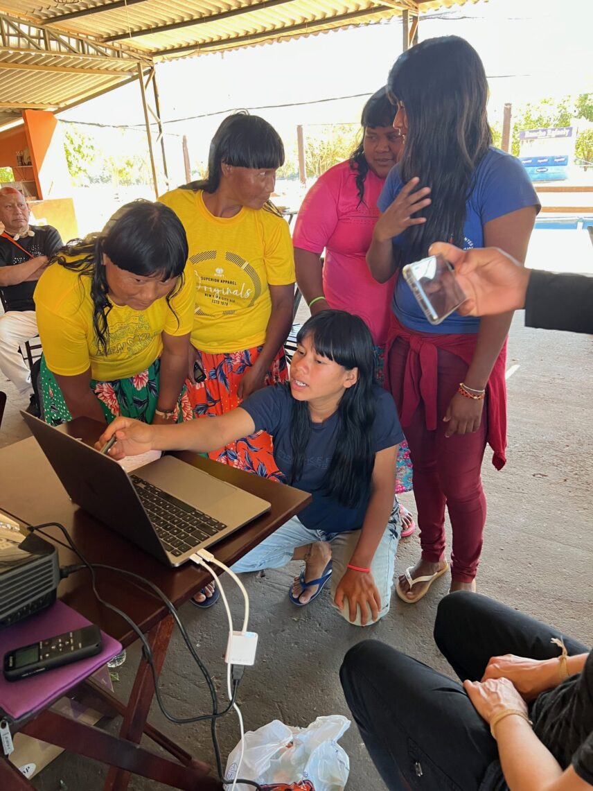 Five young A’uwẽ women crowd around a laptop with one of them pointing to the screen.