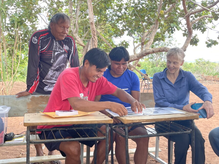 Two young A’uwẽ men and a middle-aged white woman are seated on a bench. They are smiling and arranging photographs in a binder on two small school desks. An A’uwẽ man with grey hair stands behind the bench where the three people are seated and watches them work.