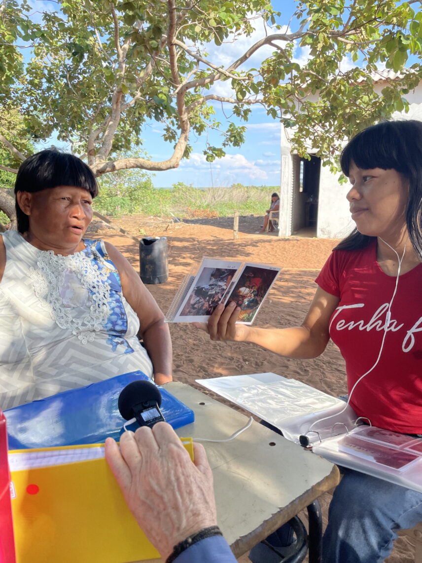 An older A’uwẽ woman sitting outdoors under a tree speaks while a younger A’uwẽ woman holds an image for her to look at with a recorder balanced on a small desk.