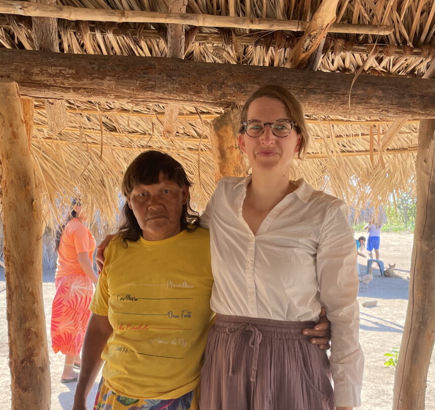 An A’uwẽ woman in a yellow t-shirt and a white woman wearing glasses and a white shirt stand with their arms around each other's shoulders with a thatched roof behind them.