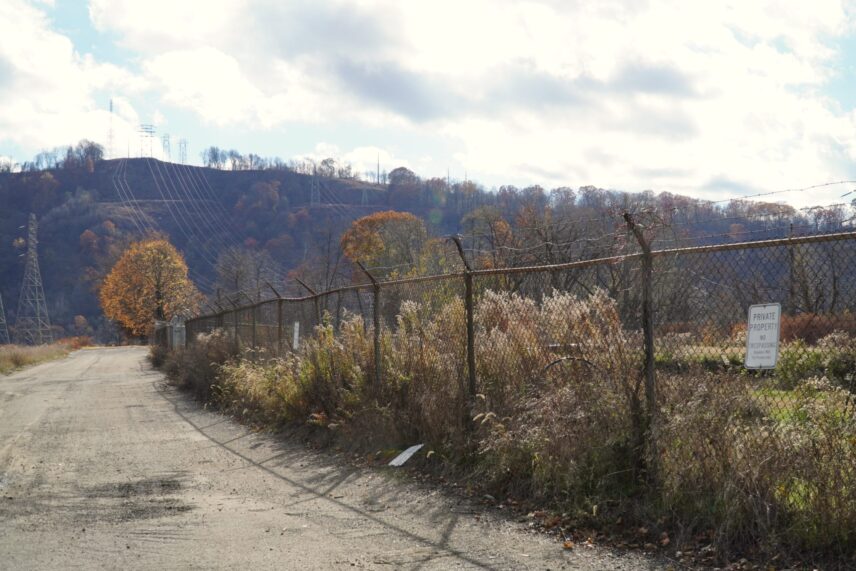 A rural gravel road runs along a chain link and barbed wire fence with power lines and tree-covered hills in the background