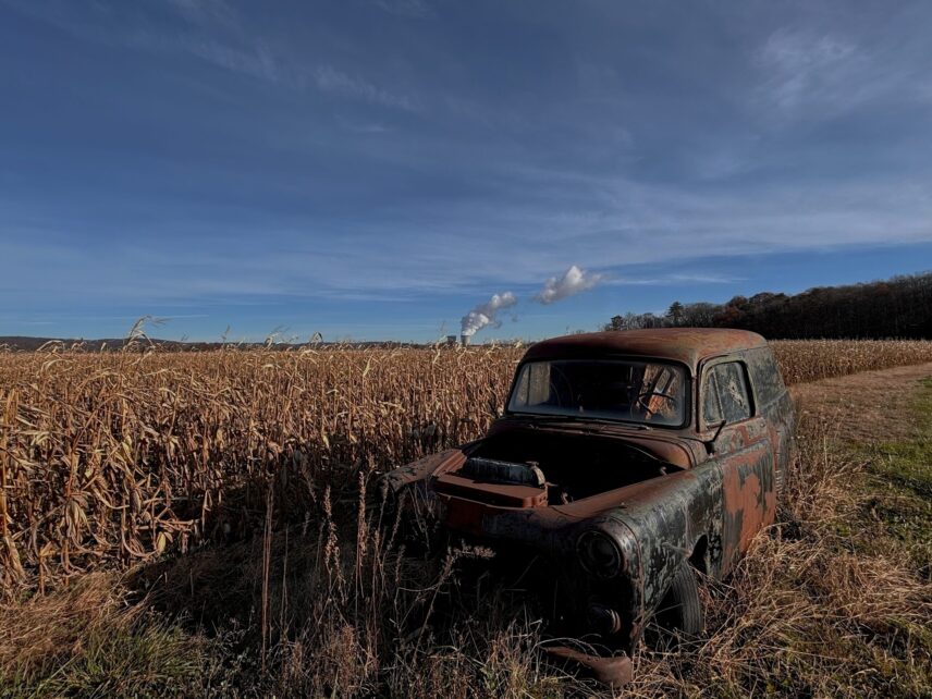 A rusted 1960s vehicle stands abandoned in a cornfield with a nuclear plant visible in the distance