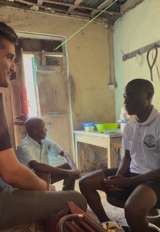 Three men sit together in a modest room in Dar es Salaam, Tanzania, speaking with one another near an open doorway.