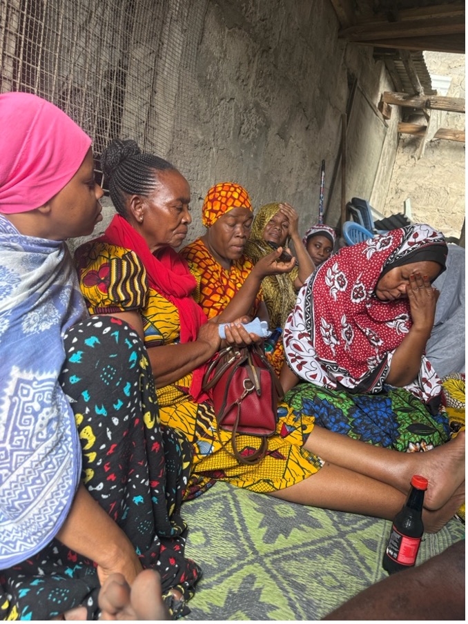 A group of women wearing colorful patterned clothing sit closely together in the hallway of a home in Buguruni Ward, Dar es Salaam, during a group discussion.