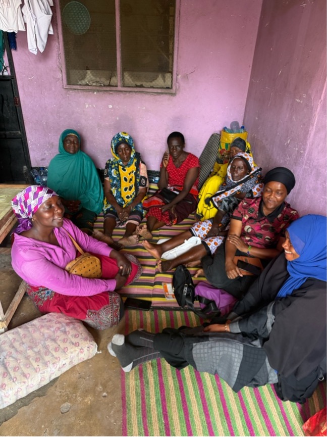 A group of women in colorful clothing and head coverings sit together on woven mats in a small room with pink walls as they gather for a widows’ group meeting in Buguruni Ward, Dar es Salaam.