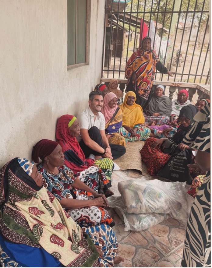 Alé Barrientos talking with a group of women in colorful patterned clothing and head coverings while they sit closely together on mats and chairs along the covered porch of a home in Buguruni Ward, Dar es Salaam, during a group discussion.