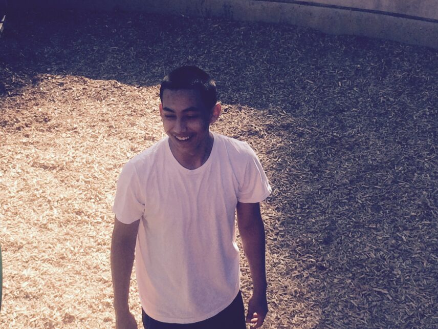 A photo that shows a teenage boy with short dark hair in a white t-shirt standing in bark mulch. He is looking down and away from the camera, and smiling.